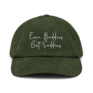 May include: A dark green corduroy baseball cap with a curved peak. The cap has the white text "Even Baddies Get Saddies" embroidered on the front. It has a structured crown and an adjustable strap at the back.