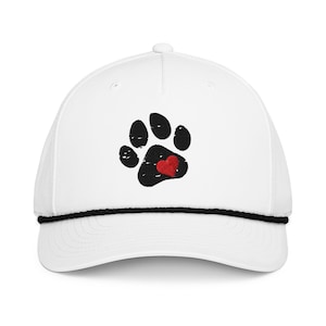 May include: White baseball cap featuring a black paw print with a red heart in the center. The cap has a black rope detail along the brim and a curved visor. The design is embroidered on the front of the cap.