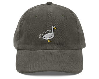May include: A corduroy baseball cap in a muted green color. The cap features an embroidered design of a white goose with a yellow beak and feet, and the words "silly goose" in a curved font.