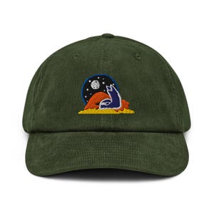 May include: A dark green corduroy baseball cap with a curved brim. The cap features an embroidered design of a blue wolf howling at the moon, set against a starry night sky and a desert landscape.