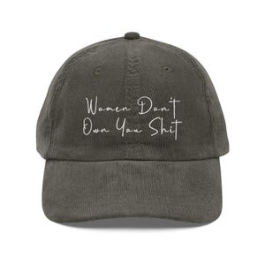 Puede incluir: Gorra de béisbol de pana verde oliva con el texto "Women Don't Own You Shit" en escritura blanca. La gorra tiene una visera curva y una correa ajustable en la parte trasera.