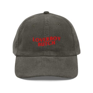 May include: A corduroy baseball cap in a muted olive green color. The cap features the embroidered text "LOVERBOY BUTCH" in a bright red color across the front. The hat has a curved brim and a rounded crown.
