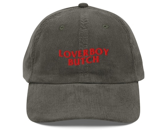 May include: A corduroy baseball cap in a muted olive green color. The cap features the embroidered text "LOVERBOY BUTCH" in a bright red color across the front. The hat has a curved brim and a rounded crown.
