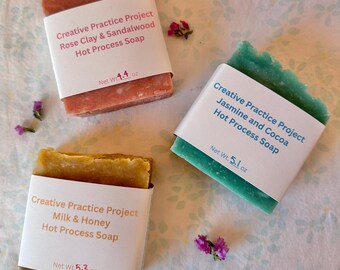 Könnte beinhalten: Drei rechteckige Seifenstücke mit weißen Papieretiketten. Eines ist Rosen-Tonerde und Sandelholz, 124,7 g; eines Jasmin und Kakao, 144,6 g; und eines Milch und Honig, 150,3 g. Jedes Etikett trägt den Text Creative Practice Project.