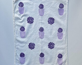 Könnte beinhalten: Ein weißes Küchentuch mit einem violetten Blumenmuster. Das Muster zeigt stilisierte Blumen in Töpfen, die sich über den Stoff wiederholen.