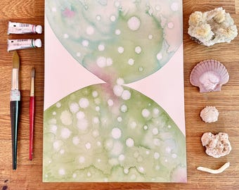 Könnte beinhalten: Aquarellmalerei in Grün und Rosa, mit weißen Punkten, in einem Sanduhr-Design. Zwei Farbtuben, zwei Pinsel und verschiedene Muscheln und Korallen sind um das Kunstwerk auf einer Holzoberfläche angeordnet.