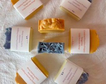 Könnte beinhalten: Sieben rechteckige Kaltseifenstücke, die in einem Kreis angeordnet sind. Vier Stücke sind orangefarben und drei sind lavendelblau. Jedes Stück hat ein weißes Papieretikett mit der Aufschrift Creative Practice Project, dem Duft und dem Nettogewicht.