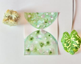 Könnte beinhalten: Ein abstraktes Aquarellgemälde in Grün- und Weißtönen, das einer Sanduhrform ähnelt, wird zusammen mit einem strukturierten, hellfarbigen Stein und einem bunten grünen Blatt gezeigt. Das Kunstwerk befindet sich auf einem weißen Hintergrund.