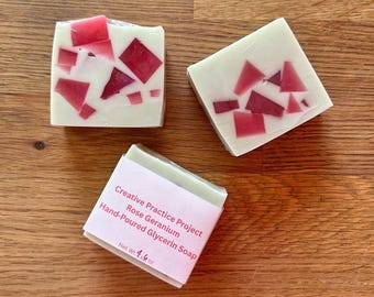 Könnte beinhalten: Drei handgemachte Glycerinseifen mit einem Mosaikdesign. Die Seife ist weiß mit roten geometrischen Formen. Eine Seife hat ein Etikett mit der Aufschrift "Creative Practice Project Rose Geranium Hand-Poured Glycerin Soap" und "Net Wt. 130 g."