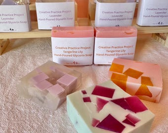 Könnte beinhalten: Eine Sammlung rechteckiger, handgegossener Glyzerinseifen von Creative Practice Project. Die Seifen enthalten eingebettete bunte geometrische Fragmente in Rosa-, Orange- und Violetttönen sowie weiße Papieretiketten mit Duftnamen wie Rose Geranium, Lavender und Tangerine Lily, einschließlich des Nettogewichts in Gramm.