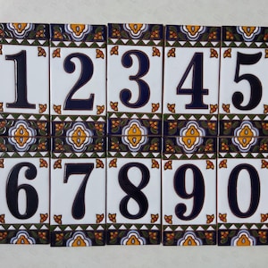May include: A set of eleven white ceramic tiles, each displaying a different number from 0 to 9, plus the number 1. The numbers are in a dark blue font. Each tile has a decorative border with floral and geometric patterns in blue, yellow, and green.