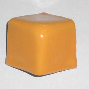 May include: A square, orange-yellow object with rounded edges. The top surface has a lighter, glossy appearance. The object is set against a plain white background, highlighting its shape and color. The overall impression is of a smooth, solid form.