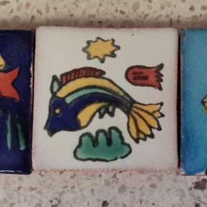 May include: Three square ceramic tiles featuring hand-painted aquatic designs. The left tile is dark blue with a multi-colored fish and green seaweed. The center tile is white with a yellow sun and a blue fish. The right tile is light blue with a red and yellow fish.