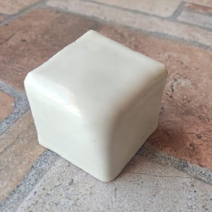 May include: A square, off-white ceramic object with rounded corners. The object is sitting on a multi-colored brick surface. The ceramic piece has a glossy finish and appears to be a decorative item.