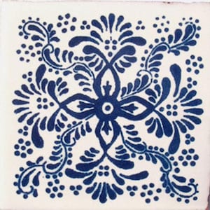 May include: A white ceramic tile with a blue floral design. The design features a central flower with four petals and intricate swirling leaves. The tile is decorated with small blue dots.