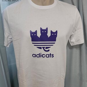 May include: White t-shirt with a purple logo of three cats in a triangle shape with the word "adicats" below.