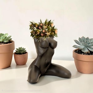 May include: A matte black ceramic torso-shaped vase featuring a kneeling human figure. The top of the vase is filled with a decorative arrangement of dried green foliage, small pink flowers, and tiny beige starfish accents.