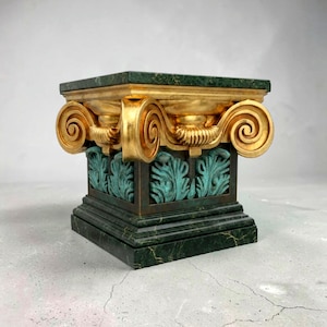 Könnte beinhalten: Ein dekorativer Sockel mit quadratischer Ober- und Unterseite, der ein goldfarbenes, verziertes Kapital mit Rollendetails aufweist. Der Sockel hat eine grün marmorartige Optik mit türkisfarbenen Akzenten und schafft eine klassische Ästhetik.