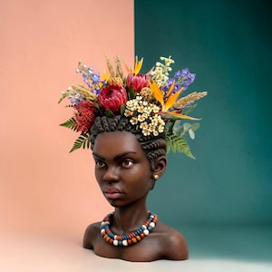 May include: A dark-skinned bust sculpture featuring braided hair adorned with a vibrant floral arrangement of red protea, orange bird of paradise, blue delphinium, and white blossoms. The figure wears a multi-layered beaded necklace in blue, white, and brown.
