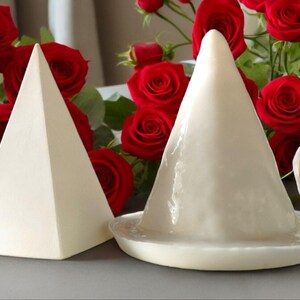 May include: Two white ceramic cake stands, one shaped like a pyramid and the other shaped like a cone, are displayed on a grey surface. The stands are surrounded by red roses.