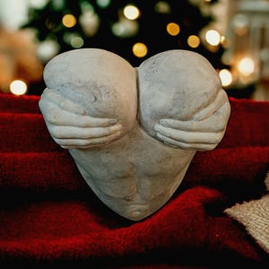 Puede incluir: Una escultura en forma de corazón de color gris claro, con manos que sostienen el corazón. La escultura está sobre un fondo rojo texturizado, con un árbol de Navidad borroso y luces en el fondo.