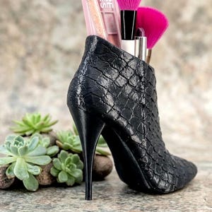 Puede incluir: Un portalápices de maquillaje negro y texturizado con forma de zapato de tacón alto. El tacón de aguja sostiene el zapato vertical, que contiene varios pinceles y aplicadores de maquillaje. Una pequeña planta suculenta está delante del zapato.