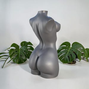 Könnte beinhalten: Eine graue, stilisierte Torsoskulptur mit glatter Oberfläche. Die Skulptur ist eine Büste, die den Oberkörper von hinten darstellt, mit definierten Kurven und anatomischen Details. Die Skulptur wird vor einem weißen Hintergrund mit grünen Pflanzen ausgestellt.