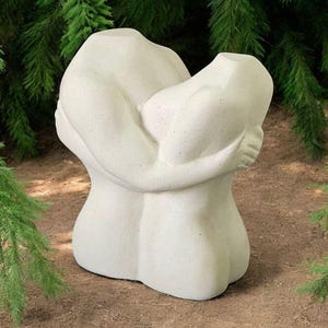 Puede incluir: Una escultura abstracta blanca de dos figuras abrazándose. La escultura está hecha de un material rugoso y texturizado y se coloca sobre un lecho de mantillo marrón.