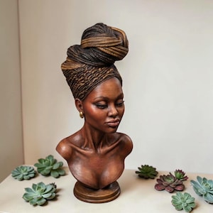 Op de afbeelding: Gedetailleerde buste sculptuur van een vrouw met een donkere huid, met een gevlochten hoofdwikkel met bruine en blonde highlights. De sculptuur heeft gouden oorbellen en staat op een houten voet. De buste is omringd door groene en paarse vetplanten.