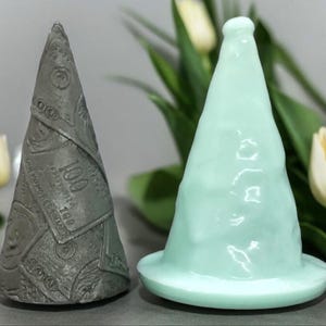 May include: Two ceramic cone-shaped candle holders. One is a light grey colour with a pattern of dollar bills. The other is a light blue colour with a smooth, textured surface.