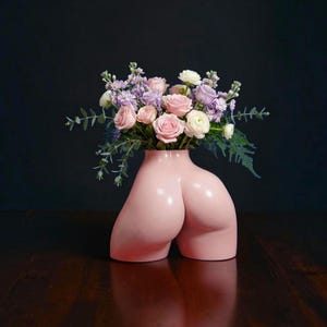 May include: A light pink ceramic vase shaped like human buttocks and hips, filled with a floral arrangement of pink roses, white ranunculus, purple stock flowers, and green eucalyptus leaves against a dark background.