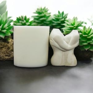 Puede incluir: Se muestra una vela cilíndrica blanca y una pequeña escultura blanquecina de figuras abrazándose. La escultura tiene un acabado liso y mate. Hay suculentas verdes en el fondo.