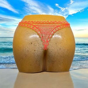 Puede incluir: Escultura de cerámica de un trasero con tanga rojo, sobre un fondo de playa y océano. La escultura es de color beige con superficie texturizada, y el tanga es de un color brillante y contrastante.