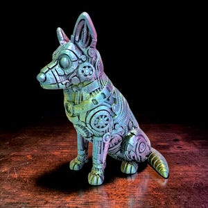 以下が含まれることがあります： スチームパンク風の、座っている犬の彫刻。犬は青、緑、紫の色合いの複雑な機械部品で構成されています。彫刻は木製の表面に置かれています。