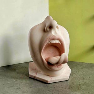 Könnte beinhalten: Eine hellfarbene, marmorartige Skulptur, die den unteren Teil eines menschlichen Gesichts darstellt, einschließlich Nase, offenem Mund und Zunge. Die Skulptur steht auf einem sechseckigen Sockel und zeigt realistische Texturen an Lippen und Zähnen.