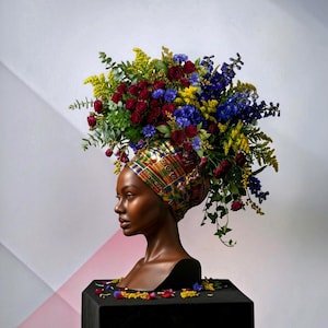 Könnte beinhalten: Eine dunkelhäutige Büste mit einem farbenfrohen, gemusterten Kopftuch, das mit einem großen, üppigen Blumenarrangement geschmückt ist. Der Strauß besteht aus dunkelroten Rosen, leuchtend gelber Goldrute, blauen Ritterspornen und grünem Blattwerk.