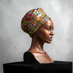 May include: A sculpture of a woman's head and shoulders, featuring a vibrant head wrap. The wrap displays a geometric design in red, yellow, green, and blue. The bust is set against a grey backdrop and sits on a black plinth.