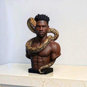 May include: A detailed sculpture bust of a dark-skinned person with short, textured hair, featuring a large, patterned brown and tan snake coiled around the neck and shoulders. The figure is shirtless, showing defined chest and abdominal muscles.