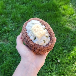 Puede incluir: Un alimento de peluche tejido a crochet con forma de patata asada con un exterior marrón y un interior blanco. La patata está cubierta con una cucharada de mantequilla de crochet blanco y amarillo.