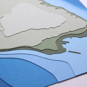 May include: A paper craft depicting a stylized island with layered blue, green, and white paper. The island is surrounded by blue water with a textured surface.