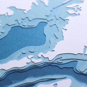 May include: A layered paper art piece featuring a topographical map design. The artwork uses shades of blue and white to depict a landscape with varying depths. The paper is cut and layered to create a 3D effect.