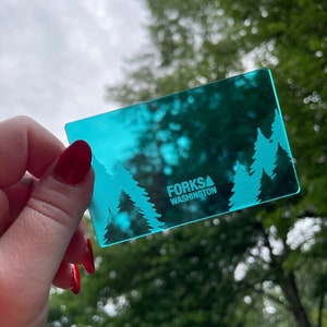 May include: A turquoise blue plastic card with the text "FORKS WASHINGTON" and a stylized image of three evergreen trees. The card is being held in a hand.