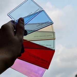 May include: A hand displays a set of colourful, transparent keyrings against a cloudy sky. Each keyring is a different colour and features text from the Twilight film series, including "twilight (2008)" and "Breaking Dawn Part 2 (2012)."