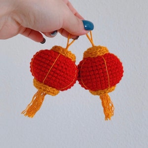 May include: Two red crocheted Chinese lanterns with gold trim and tassels. The lanterns are hanging from a string.