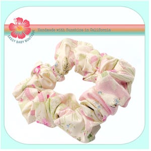 Puede incluir: Una scrunchie hecha a mano con un diseño de patchwork. La tela presenta una mezcla de colores rosa, verde y crema con estampados florales y geométricos. El texto "Sassy Baby Boutique" y "Handmade with Sunshine in California" son visibles.