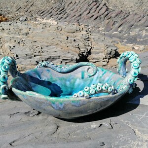 May include: A ceramic bowl with octopus tentacles for handles. The bowl is glazed in shades of blue and green, with a white and gray speckled finish.