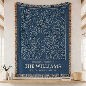 May include: A navy blue woven throw blanket featuring a white topographic map design, the text GATLINBURG, TENNESSEE, THE WILLIAMS, and the coordinates 35°42'51"N 83°30'37"W EST. 2026. The blanket has a decorative fringe border with multicolored threads.