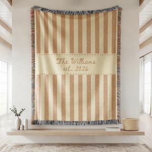 Puede incluir: Una manta a rayas beige y marr&oacute;n con el texto "The Williams est. 2026". La manta tiene un borde con flecos y est&aacute; colgada en una pared. La manta es un art&iacute;culo decorativo para el hogar.