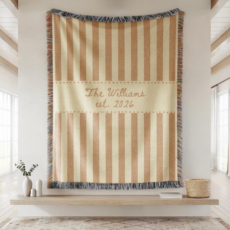 Puede incluir: Una manta a rayas beige y marr&oacute;n con el texto "The Williams est. 2026". La manta tiene un borde con flecos y est&aacute; colgada en una pared. La manta es un art&iacute;culo decorativo para el hogar.