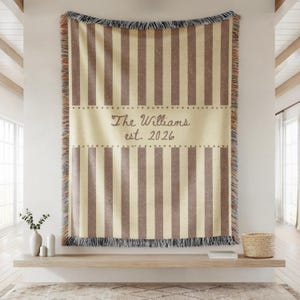Puede incluir: Una manta a rayas beige y marr&oacute;n con un borde con flecos. La manta presenta el texto "The Williams est. 2026" en una fuente decorativa. La manta se exhibe en una estanter&iacute;a de madera.